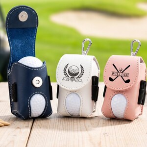 May include: Three golf ball pouches in navy blue, white, and light pink. Each pouch features a magnetic snap closure, a metal carabiner, and side loops for tees. The white pouch displays a golf ball emblem with the name Hayden, and the pink pouch shows crossed golf clubs with the name William.