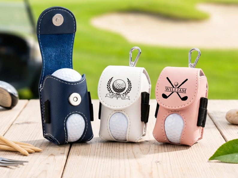 May include: Three golf ball pouches in navy blue, white, and light pink. Each pouch features a magnetic snap closure, a metal carabiner, and side loops for tees. The white pouch displays a golf ball emblem with the name Hayden, and the pink pouch shows crossed golf clubs with the name William.