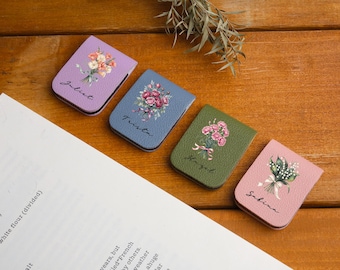 May include: Four leather book page holders in lavender, blue, olive green, and pink. Each features a floral bouquet design and a name: Juliet, Trista, Hazel, and Sabina. The items are on a wooden surface.