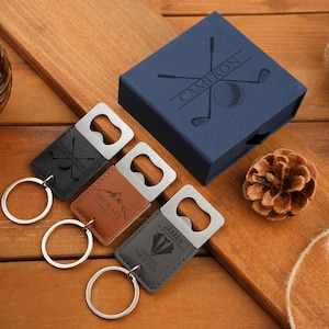 May include: A collection of personalized bottle opener keychains with silver metal openers and leatherette covers. The keychains are black, brown, and gray, with engraved golf-themed designs and names. A navy blue gift box with a golf ball and club design is also present.
