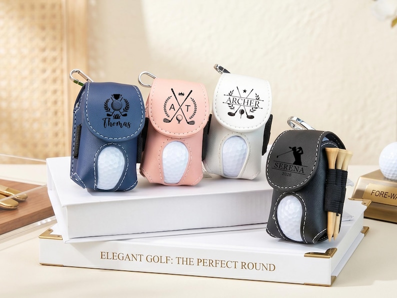 May include: Four golf ball pouches in navy blue, light pink, white, and black, each featuring a metal clip, a cutout for a golf ball, and personalized engravings. The black pouch includes three wooden golf tees attached to the side.