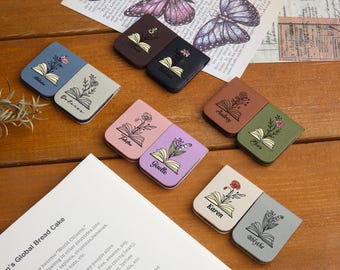 May include: A collection of personalized leather book page markers in various colors, each featuring a floral design and a name. The markers are arranged on a wooden surface, with a book and decorative paper in the background.