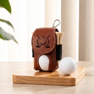 May include: A brown leather golf ball pouch with a flap closure featuring a black engraved logo of two crossed golf clubs and a ball with the letters TN. The pouch holds wooden golf tees and has a metal clip, with a white golf ball placed beside it.