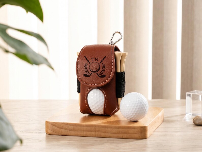 May include: A brown leather golf ball pouch with a flap closure featuring a black engraved logo of two crossed golf clubs and a ball with the letters TN. The pouch holds wooden golf tees and has a metal clip, with a white golf ball placed beside it.