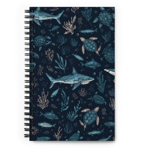 May include: Spiral-bound notebook featuring a dark blue background with an all-over pattern of marine life. The design includes detailed illustrations of sharks, sea turtles, fish, and seaweed in shades of blue and beige. The notebook's spiral binding is black.