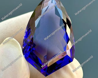 May include: A faceted, teardrop-shaped gemstone with a deep blue and purple color gradient. The gem is held by a gloved hand, with the text "Maheragemsworld" repeated around the edges.