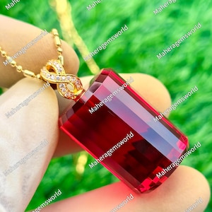 May include: A rectangular, deep red gemstone pendant suspended from a gold-toned chain. The pendant features a gold-toned infinity symbol bail encrusted with small, clear, sparkling stones, connecting the gemstone to the delicate ball chain.