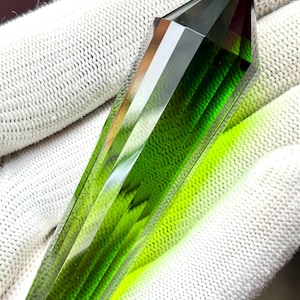May include: A faceted, elongated gemstone with a pointed tip. The gem transitions from a deep green at the top to a lighter, yellowish-green towards the base. The facets create a play of light and shadow, enhancing its crystalline structure.