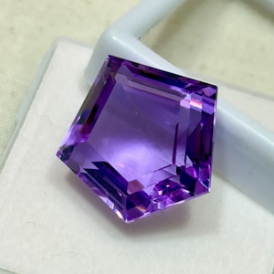 Peut inclure: Une pierre précieuse d'améthyste taillée en forme de pentagone. La gemme est d'un violet profond avec de multiples facettes qui réfléchissent la lumière. Elle est posée sur un fond blanc et un support en plastique blanc.