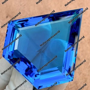 May include: A large, faceted gemstone with a striking blue hue. The gem is cut into a unique kite shape, showcasing multiple facets that reflect light. The text "Maheragemsworld" is repeated around the edges of the gem.