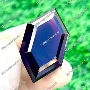 Könnte beinhalten: Ein facettierter, geometrischer Edelstein mit tiefvioletter Farbe und polierter Oberfläche. Der Edelstein wird vor einem verschwommenen grünen Hintergrund gehalten. Der Text "Maheragemsworld" wiederholt sich am Rand des Bildes.