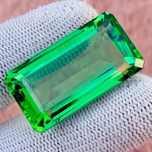 May include: A vibrant, rectangular-cut, emerald-green gemstone. The gem is faceted with sharp edges and a polished surface, reflecting light. The stone is held by a white glove, highlighting its clarity and color.
