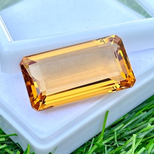 Op de afbeelding: Een rechthoekige, gefacetteerde edelsteen met een warme, goudgele tint. De edelsteen is geplaatst tegen een witte achtergrond, met groen gras op de achtergrond. De slijping van de steen toont zijn helderheid en schittering.