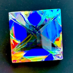 May include: A square, multi-colored glass prism with a faceted design. The prism features vibrant hues of blue, yellow, red, and green, creating a kaleidoscopic effect. The object is set against a dark background.
