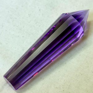 May include: A faceted, elongated crystal in shades of purple. The crystal has a pointed tip and a flat base. The facets create a striped effect, reflecting light. The crystal is likely a decorative item or a gemstone.