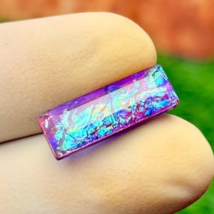 May include: A rectangular cabochon featuring a deep purple base with an iridescent, multi-colored foil inlay that reflects shades of blue, green, and magenta. The surface is smooth and polished, showcasing a vibrant, shimmering, and reflective texture.