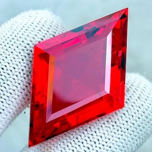 May include: A vibrant, faceted, red gemstone with a unique kite shape. The gemstone is held by a white glove, showcasing its brilliant cut and intense color. The gem's facets reflect light, creating a dazzling display.