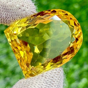 Könnte beinhalten: Ein großer, facettierter Edelstein in leuchtendem Gelb, gehalten in einer behandschuhten Hand. Der Schliff des Edelsteins zeigt zahlreiche reflektierende Oberflächen, die ein schillerndes Spiel von Licht und Farbe erzeugen. Der Hintergrund ist ein sanftes Grün.