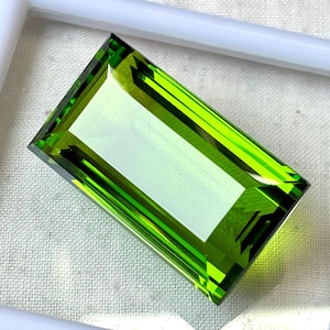 May include: A rectangular, faceted gemstone with a vibrant, lime green hue. The gem is set within a white frame, showcasing its sharp angles and internal reflections. The stone's clarity and color suggest a high-quality cut.