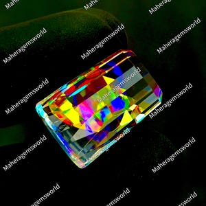 May include: A rectangular faceted gemstone displaying a vibrant spectrum of iridescent colors including red, yellow, blue, and green. The stone features a clear, highly reflective surface with sharp geometric cuts that create a prismatic effect.