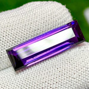 May include: A rectangular, faceted gemstone in a deep purple shade. The gem is long and slender, featuring a lighter central band. The edges are sharply defined, and the stone appears polished, reflecting light.