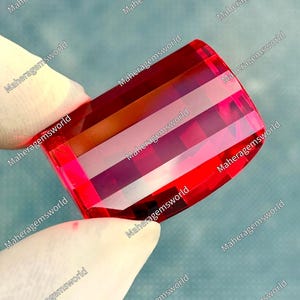 May include: A rectangular, faceted gemstone with a vibrant, translucent red colour. The stone features a step-cut design with horizontal facets that create a layered, geometric appearance. The surface is polished and reflects light clearly.