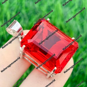 Könnte beinhalten: Ein großer, facettierter, rechteckiger roter Edelsteinanhänger mit einer silberfarbenen Öse. Der Edelstein ist in einer silbernen Fassung gehalten. Der Hintergrund ist ein verschwommenes Grün. Der Text "Maheragemsworld" wiederholt sich im Bild.