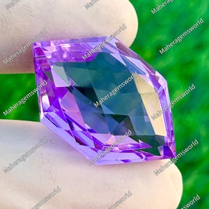 May include: A faceted, purple gemstone with multiple reflective surfaces. The gem is held by a gloved hand, with the text "Maheragemsworld" repeated around the edges. The gem's cut creates a play of light and shadow.