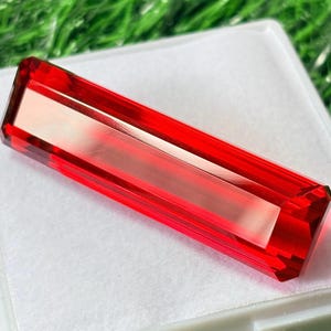 May include: A rectangular, faceted, red gemstone with a clear, rectangular center. The gemstone is displayed on a white surface, with green grass visible in the background. The gemstone's edges are sharply defined, and the color is a vibrant, transparent red.