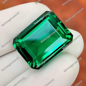 May include: A vibrant, emerald-green gemstone with a rectangular step-cut facet pattern. The stone displays high clarity and deep green saturation, featuring sharp, clean edges and a polished surface that reflects light across its geometric facets.