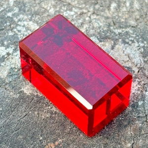 Può includere: Un blocco rettangolare di vetro trasparente rosso rubino con bordi smussati. Il blocco è appoggiato su una superficie ruvida e grigia, mostrando il suo colore vibrante e la forma geometrica. La luce cattura le sfaccettature.