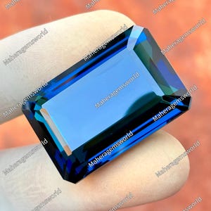 Puede incluir: Una gema rectangular de color azul intenso con múltiples facetas, sostenida por una mano enguantada. El texto "Maheragemsworld" se repite alrededor de los bordes de la imagen.