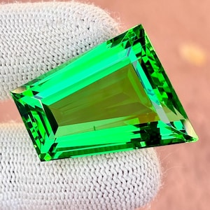 May include: A vibrant, emerald-green gemstone with a rectangular shape and angled corners. The gem is displayed against a white knitted fabric, highlighting its clarity and colour. The cut of the stone creates multiple reflective surfaces.