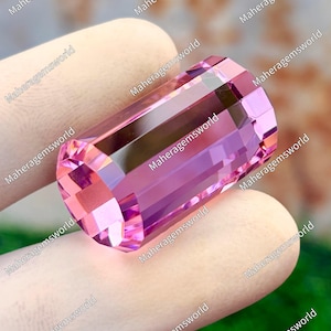 May include: A faceted, rectangular pink gemstone. The gem has a light pink hue with a darker pink interior. The edges are sharply cut, reflecting light. The text "Maheragemsworld" is repeated around the image.