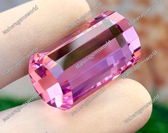 Puede incluir: Una gema rosa rectangular y facetada. La gema tiene un tono rosa claro con un interior rosa más oscuro. Los bordes están afilados, reflejando la luz. El texto "Maheragemsworld" se repite alrededor de la imagen.