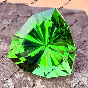 May include: A bright green, faceted gemstone with a geometric design. The gem is cut with multiple facets that reflect light, creating a sparkling effect. The gemstone is displayed on a rough, textured surface.