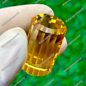 Puede incluir: Una gema facetada de color amarillo dorado sostenida por una mano enguantada. La gema tiene forma cilíndrica con múltiples facetas, que reflejan la luz. El fondo es verde borroso, con el texto "Maheragemsworld" repetido.