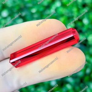 May include: A vibrant red, elongated, faceted gemstone with a rectangular, bar-like shape. The crystal features sharp, clean edges and a polished, reflective surface that catches light, displaying a deep, uniform crimson hue throughout its structure.