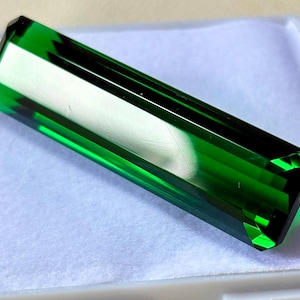 May include: A rectangular, faceted, emerald-green gemstone. The gem has a polished surface with sharp, defined edges. The color is a deep, rich green, with a slight transparency. The gemstone is set against a white background.