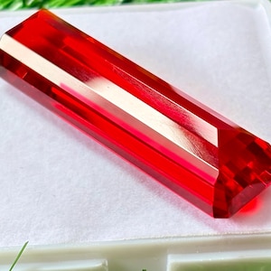 May include: A faceted, rectangular gemstone with a vibrant red hue. The gem has a polished, reflective surface and a geometric design. The gemstone is displayed on a white surface.