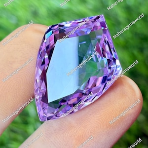 May include: A striking, shield-cut gemstone in shades of purple, with a central, lighter facet. The gem is held between fingers, highlighting its intricate facets and reflective qualities. The background is a blurred green.