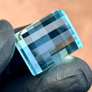 May include: A rectangular, faceted gemstone with a light blue hue. The gem is held by a gloved hand, showcasing its intricate, geometric cuts. The facets create a play of light and shadow, highlighting the gem's clarity and brilliance.