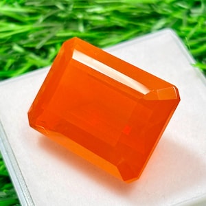 Puede incluir: Una gema rectangular de color naranja brillante con un corte facetado. La gema se muestra sobre una superficie blanca, con un fondo verde borroso. El color de la gema es un naranja rico y saturado, y sus facetas captan la luz.