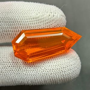 May include: A faceted, orange gemstone with a rectangular shape and angled ends. The gem is held against a white, knitted fabric, showcasing its vibrant colour and intricate cuts. The gemstone's facets reflect light, creating a sparkling effect.