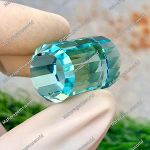 May include: A faceted, cylindrical gemstone with a light blue hue. The gem is held by a gloved hand, showcasing its geometric cuts and polished surface. The background is blurred, emphasizing the gem's clarity and color.