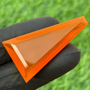 Puede incluir: Una gema geométrica de color naranja brillante con forma triangular. La gema tiene una superficie pulida y facetada que refleja la luz, creando una exhibición brillante. La gema se sostiene contra una superficie negra con un fondo verde.