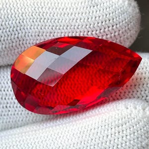 Può includere: Una gemma rossa sfaccettata a forma di goccia. La gemma ha una superficie brillante e riflettente con molteplici sfaccettature che catturano la luce. La pietra è posta su uno sfondo bianco strutturato.