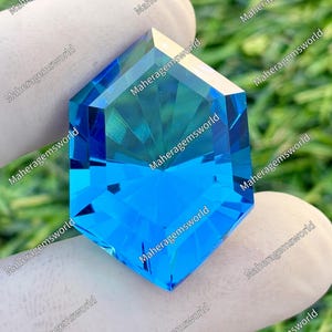 Op de afbeelding: Een gefacetteerde, zeshoekige edelsteen in blauwtinten, vastgehouden door een gehandschoende hand. De edelsteen heeft een briljante slijpvorm, die licht reflecteert en de helderheid laat zien. De achtergrond is wazig groen.