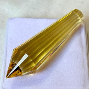 Puede incluir: Una punta de cristal amarillo dorado facetada. La gema es alargada con una punta y una base cilíndrica plana. El cristal se muestra sobre una superficie rectangular blanca.