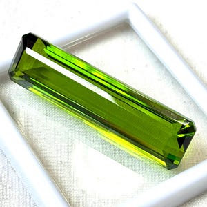 May include: A rectangular, faceted gemstone with a vibrant, lime green hue. The gem is set within a white, square frame, highlighting its clarity and brilliance. The cut of the stone allows light to reflect, creating a captivating display.